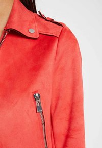 Czerwona kurtka z sztucznego zamszu z klapami, srebrnymi detalami i asymetrycznym zamkiem po lewej stronie. Miękka faktura i gładkie wykończenie.