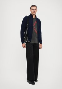 Cárdigan de cremallera navy sobre una camiseta gris con texto rojo, combinado con pantalones de pierna ancha negros y zapatos negros. Texturas oscuras y suaves.