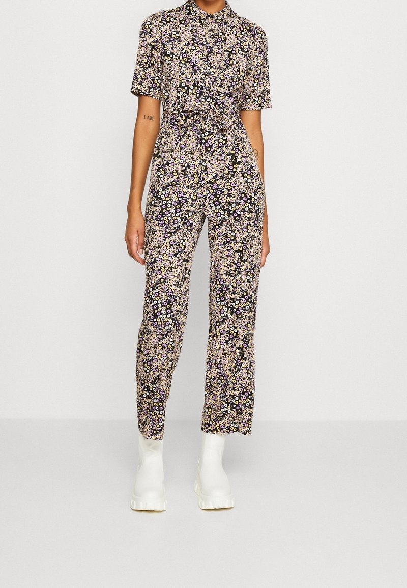 Combinaison florale à manches courtes, fond noir, petites fleurs multicolores, taille ceinturée et jambes ajustées. Associée à des bottes blanches à semelles épaisses.