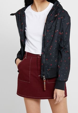 Kobieta ubrana w biały krótki t-shirt, czarną kurtkę na zamek z kwiatowym wzorem oraz bordową spódnicę z wysokim stanem i przednimi kieszeniami.