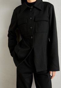 Czarna zapinana na guziki kurtka wykonana z miękkiego materiału, z dwoma kieszeniami na piersi, ostro zakończonym kołnierzem i długimi rękawami o luźnym kroju.