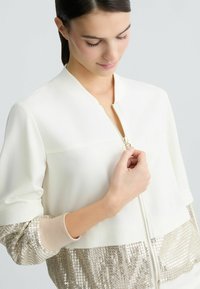 White bomber jacket with a zip front, featuring shiny gold sequin accents on the lower half and textured sleeves. Smooth fabric, relaxed fit.