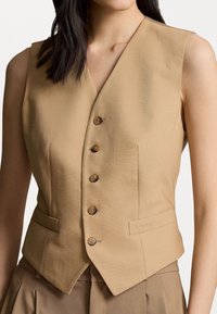 Béžová vesta s výstřihem do V, se šesti knoflíky na zapínání, dvěma bočními kapsami a na míru střiženým střihem. Vyrobena z texturované látky.