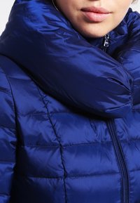 Marine blauwe pufferjas met een hoge kraag, voorzien van een ritssluiting en een gewatteerde structuur. Het materiaal is glad en glanzend.