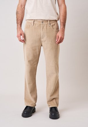 Man met beige ribfluwelen broek, zwarte schoenen en een lichtbeige overhemd staand tegen een effen achtergrond.