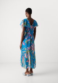 Abito blu floreale fluido con maniche a volant, schiena a V profonda e tessuto trasparente. Decorato con fiori rosa e verdi. Modello che indossa sandali con tacco azzurri chiari.