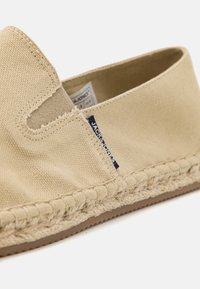 Beige kanvas slipsko med elastiske sideåbninger, flettet jute-detalje langs sålen og et lille blåt mærkat.