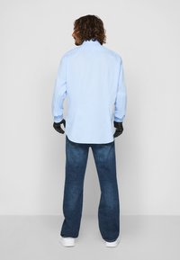 Lyseblå skjorte med lange ermer, avslappet passform. Sammen med mørkeblå jeans og hvite joggesko. Hender i svarte hansker synlige.