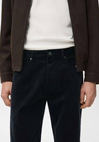 Pantalones oscuros de pana con superficie texturizada, con cierre de botón y trabillas para cinturón. Combinados con una camisa blanca y una chaqueta marrón con cremallera.