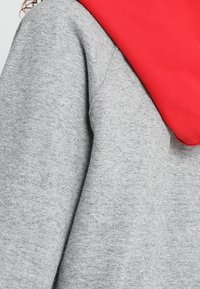 Sudadera gris con capucha roja. La tela parece suave y texturizada. El diseño presenta un corte casual y atlético.