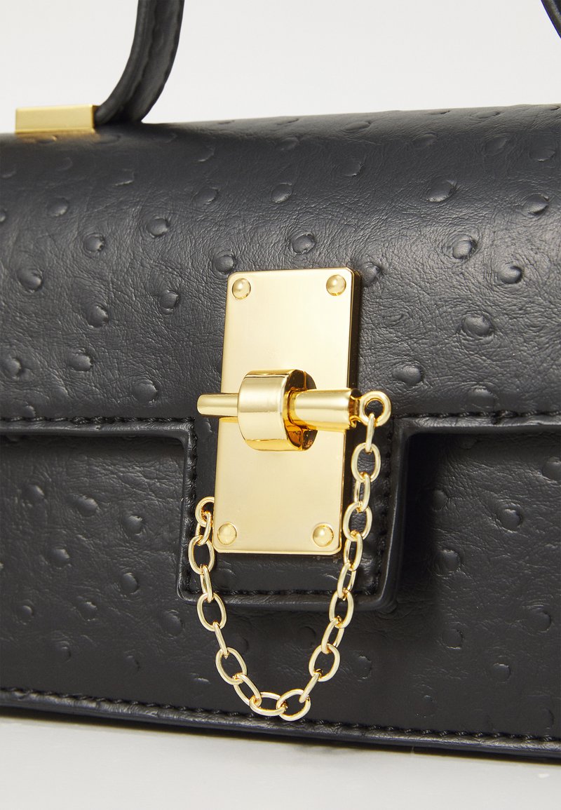 Sac à main en cuir noir avec un motif en relief, quincaillerie dorée et accent en chaîne. Présente une forme structurée et une fermeture rectangulaire.