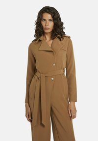 Bruine trenchcoat met een brede kraag, dubbele knopen en een ceintuur in de taille. Heeft lange mouwen en een gladde, gestructureerde stof.