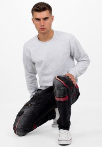 Sweat-shirt gris clair, jeans noirs avec des accents rouges et des fermetures éclair, portés avec des baskets blanches. Les jeans présentent plusieurs poches et un design ajusté.