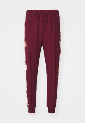 Burgundy joggers made from soft fabric, featuring white side stripes and an embroidered logo patch on the left thigh. Elastic waistband with drawstring.