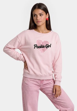 Giovane donna che indossa una felpa rosa con un cuore e il testo "Pookie Girl", pantaloni rosa e orecchini rossi a forma di cuore, posando con la mano sul fianco.