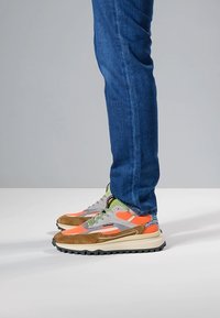 Sneakers sportive in marrone, arancione e grigio con tomaia in mesh e pannelli testurizzati, caratterizzate da una suola bianca spessa e accenti vivaci.