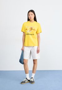 Yellow graphic t-shirt with "Cyclone" print, paired with white striped shorts and black sneakers. Model holds a denim item. Blue backdrop.
