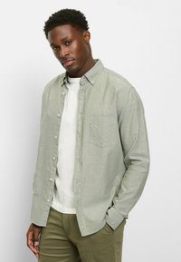 Hellgrünes Hemd zum Knöpfen mit Brusttasche, Kragen und langen Ärmeln, getragen über einem weißen T-Shirt und olivgrünen Hosen.