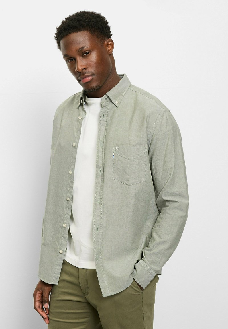 Hellgrünes Hemd zum Knöpfen mit Brusttasche, Kragen und langen Ärmeln, getragen über einem weißen T-Shirt und olivgrünen Hosen.