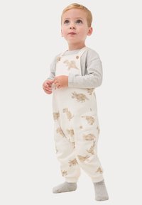 Weiße Latzhose mit beigen Dinosaurierdruck, Holzknoten-Details und Seitentaschen. Kombiniert mit einem grauen Langarmshirt und grauen Socken.