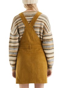 Mosterdkleurige corduroy tuinbroekjurk met een getailleerde taille en achterzakken, gecombineerd met een gebreide trui met meerkleurige strepen.