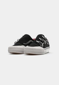 Zapatillas negras sin cordones con herrajes plateados, suela de goma blanca texturizada y parte superior lisa mate. Presentan acentos decorativos de cadena.