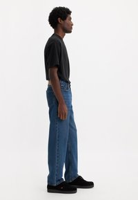 T-shirt noir à manches courtes, jean bleu à jambes droites, et chaussures en suède noir. Coupe slim avec texture denim ; le jean présente des coutures visibles et des rivets.