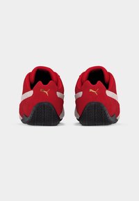 Ζευγάρι κόκκινων αθλητικών παπουτσιών Puma, φωτογραφημένα από πίσω, με λευκές ρίγες και μαύρες υφασμάτινες σόλες σε απλό φόντο.