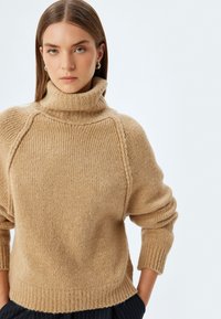 Maglione beige lavorato a maglia con colletto alto, texture a costine e maniche a sbuffo; presenta cuciture raglan e una silhouette rilassata.