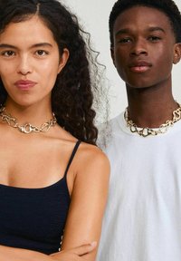 Collar de cadena de oro con formas de eslabones abstractos, llevado por una persona con cabello rizado y otra que viste una camisa blanca lisa.