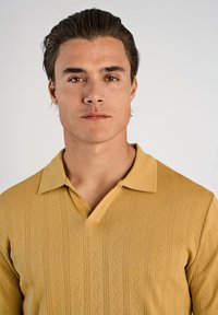 Musztardowożółta koszulka polo o strukturze tkaniny i kołnierzyku z wycięciem. Posiada pionowe wzory w paski oraz krótkie rękawy. Gładka tkanina.