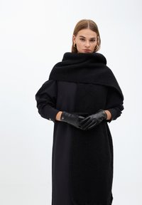 Černý pletený šál se přesně překlápí přes černé šaty se saténovým povrchem. Modelka má na sobě černé prošívané rukavice se zlatými akcenty, které představují minimalistický design.