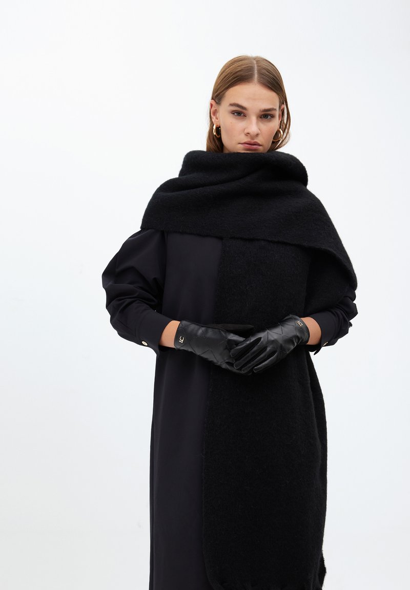 Černý pletený šál se přesně překlápí přes černé šaty se saténovým povrchem. Modelka má na sobě černé prošívané rukavice se zlatými akcenty, které představují minimalistický design.