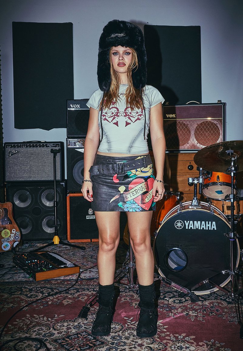 Sombrero de pelo sintético negro, camisa ajustada de color azul claro con gráfico rojo, falda corta de cuero negro con estampados coloridos y botas negras con cordones.