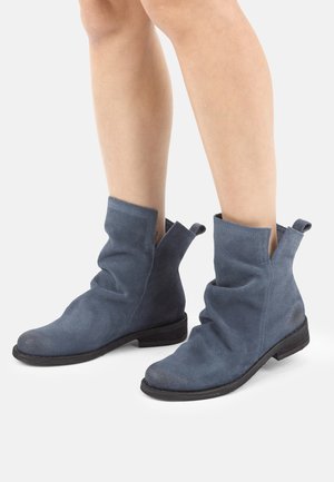 Gambe con stivaletti alla caviglia in camoscio blu dal design morbido e suole piatte nere, in piedi su uno sfondo bianco semplice.