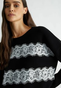 Maglione nero con due strisce di pizzo orizzontali che presentano motivi floreali e bordi frangiati, progettato con un collo a giro e una vestibilità morbida.