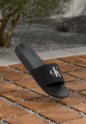 Črne natikače z širokim trakom, ki ima bel logotip. Mehki material, ravna: podplat in minimalistična zasnova. Teksturna oblika na podplatu.