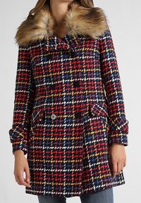 Dvouřadový kabát z vlněné směsi s červeným, modrým, žlutým a bílým houndstooth vzorem, s odnímatelným límcem z umělé kožešiny.