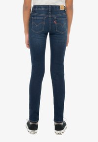 Tumed sinised kitsad teksapüksid, mis on valmistatud denimist, kõrge vöökohaga, tagataskutega, millel on kõrgendatud õmblused, ning tagaküljel beežist nahast brändimärk.