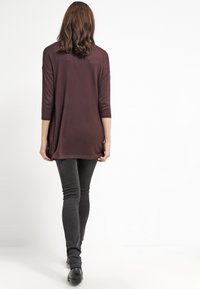 Žena stojící zády, oblečená v volném tmavě fialovém topu, přiléhavých černých džínách a černých botách na pozadí s čistě bílou barvou.