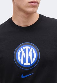 Κοντινό πλάνο ενός ατόμου που φοράει μαύρο t-shirt με μπλε και λευκό κυκλικό λογότυπο και μπλε σήμα Nike από κάτω.