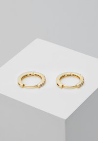 Goldene Creolen mit einer glatten Oberfläche, die entlang einer Kante eine Reihe kleiner klarer Steine aufweisen. Kreisform, zeitgenössisches Design.