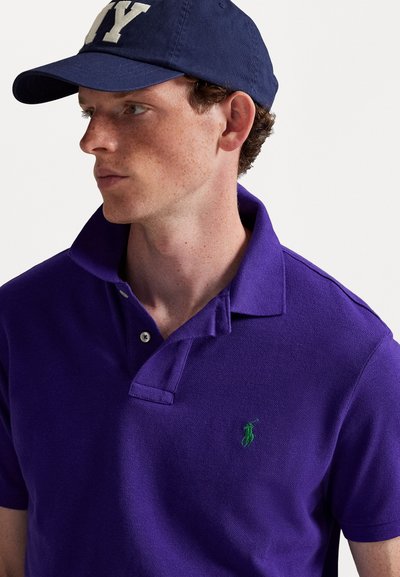 Camisa polo roxa com um tecido texturizado e um logotipo verde bordado. A camisa apresenta um colarinho clássico e uma pala com dois botões.