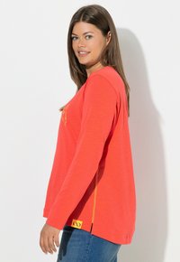 Langärmeliges, korallenoranges Hemd mit gelben Akzenten, lockerer Passform, seitlichen Schlitzen und Logo am Saum. Weicher Stoff mit glatter Textur.