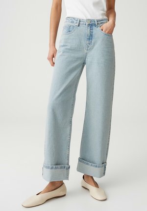 Donna che indossa jeans a gamba larga color azzurro chiaro con risvolti, top bianco e scarpe basse color crema, in piedi con una mano in tasca.