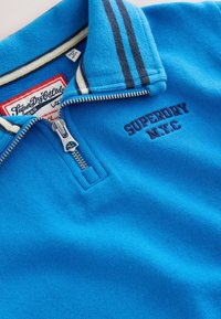 Pile blu con colletto a zip, decorato con la scritta ricamata "SUPERDRY N.Y.C" in blu scuro e dettagli a righe in blu navy e crema.