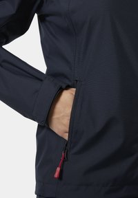 Marineblauw jack met een gestructureerde stof, voorzien van een zijzak met ritssluiting en een rood trekkoord. Zichtbaar manchetdetail met een nauwsluitend design.