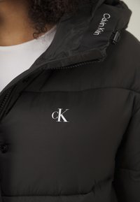 Juodas pūkų švarkas su lygiu paviršiumi, turintis užsegimą su mygtukais ir "CK" logotipu. Su kapišonu, kurio tvirtinimai reguliuojami.