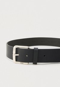 Juodas odinis diržas su sidabro spalvos stačiakampiu sagtimi, išgraviruotas "HUGO", pateiktas suvyniotas ant vientiso šviesaus fono.