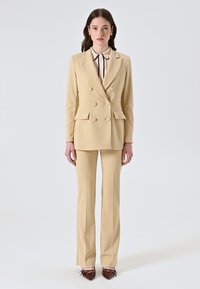 Completo beige doppiopetto con vestibilità sartoriale, dotato di due tasche anteriori e uno stile di pantaloni abbinato, abbinato a tacchi a punta marroni.